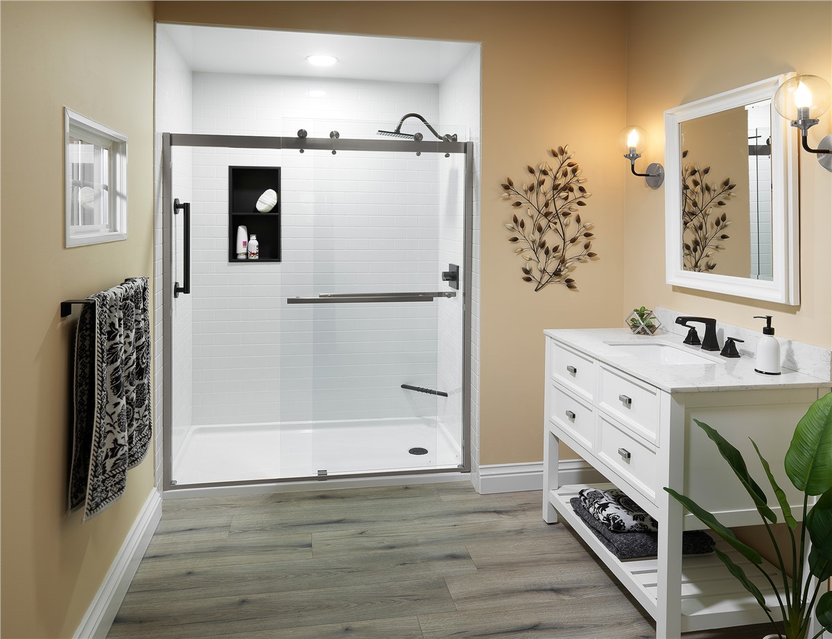 A modern bathroom with tan walls, a large white shower with glass doors, a white vanity with a mirror, two wall sconces, a towel rack, wall art, and a green plant on wood-style flooring.