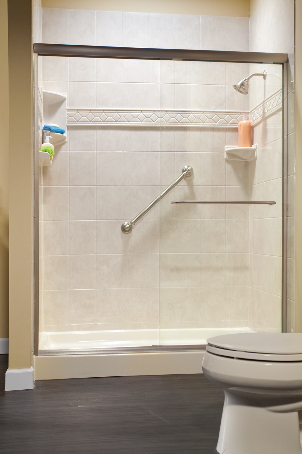 A walk-in shower with beige tiled walls, a metal grab bar, built-in shelves holding bottles, a mounted showerhead, and a partial view of a white toilet in the foreground. The floor appears to be dark wood or laminate.