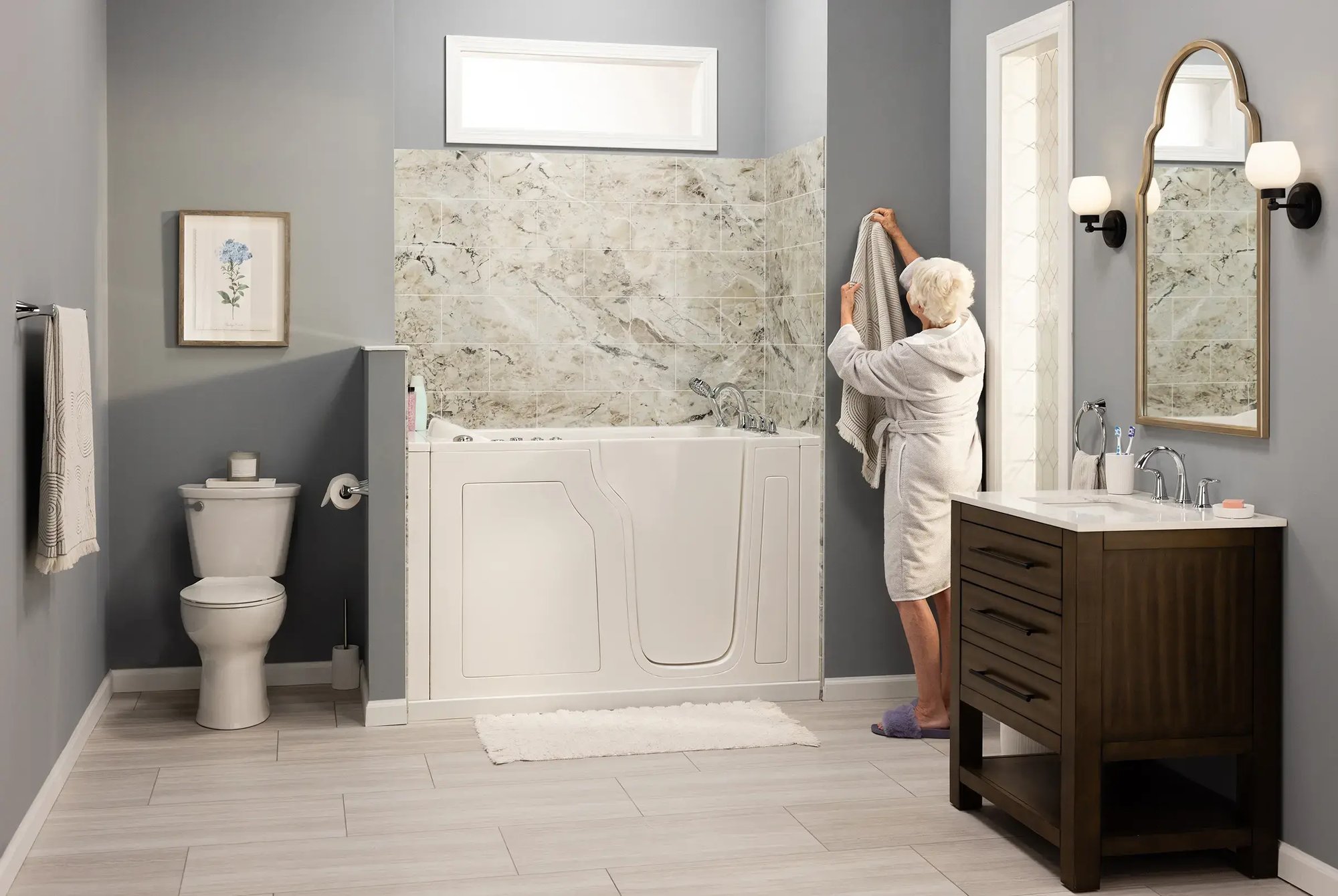 Bathroom featuring a walk-in bathtub with marble-patterned tile surround, vanity with dark brown wood cabinet, toilet, and grey walls. An elderly woman in a robe stands near the tub, holding a towel.