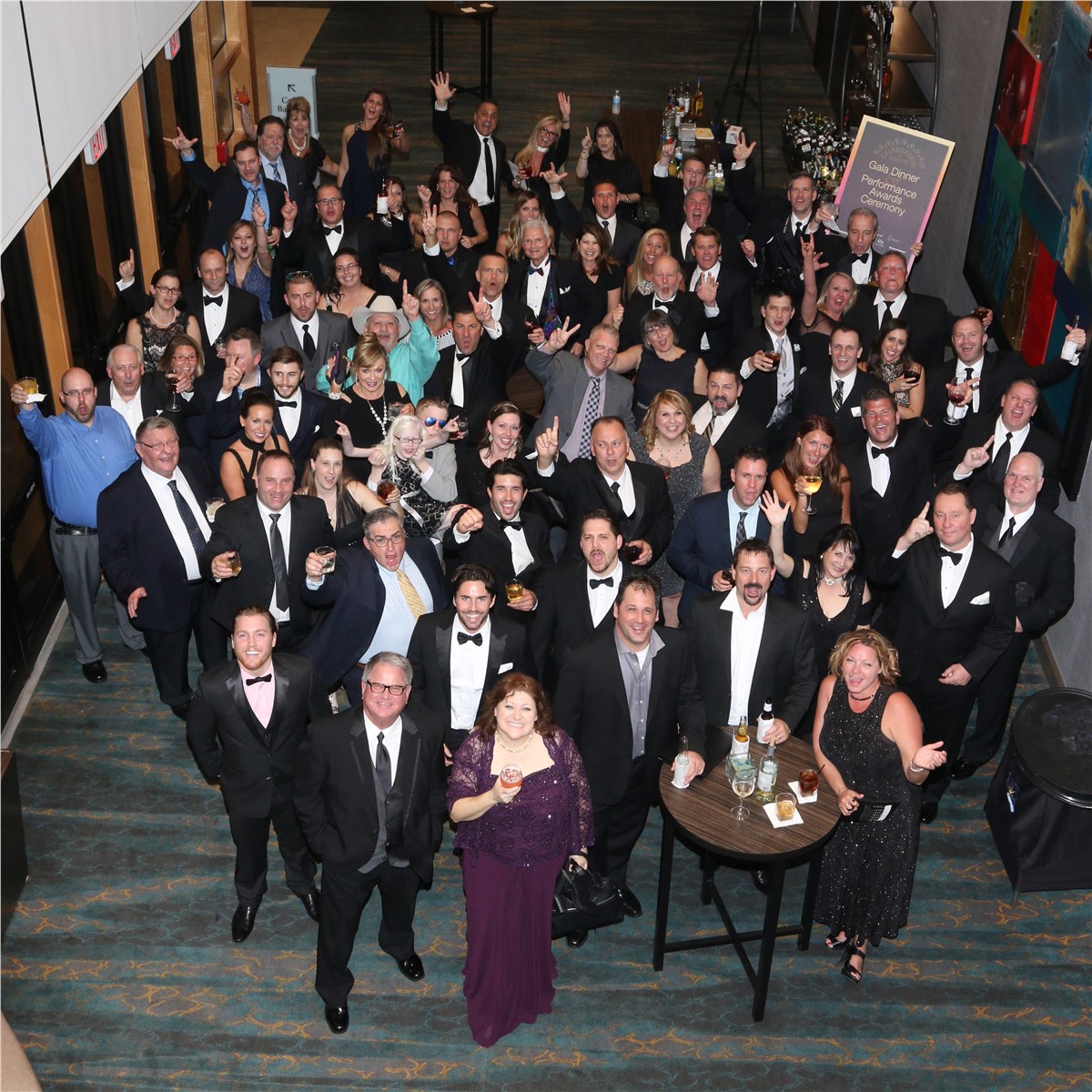 Aerial view of a large group of people in formal wear, mainly tuxedos and gowns, smiling and waving at the camera during a gala dinner performance awards ceremony.