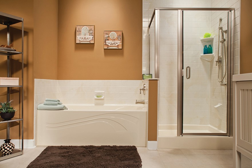 Bathroom interior featuring a large soaking tub with a tiled backsplash, adjacent to a glass-enclosed shower with a built-in shelf containing bath products. A dark brown rug sits on the tile floor in front of the tub, and the walls are painted a warm tan color with decorative artwork.