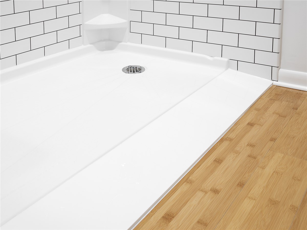 Close-up of a white shower base with a metal drain, installed in a bathroom with white subway tiles and light wood flooring.