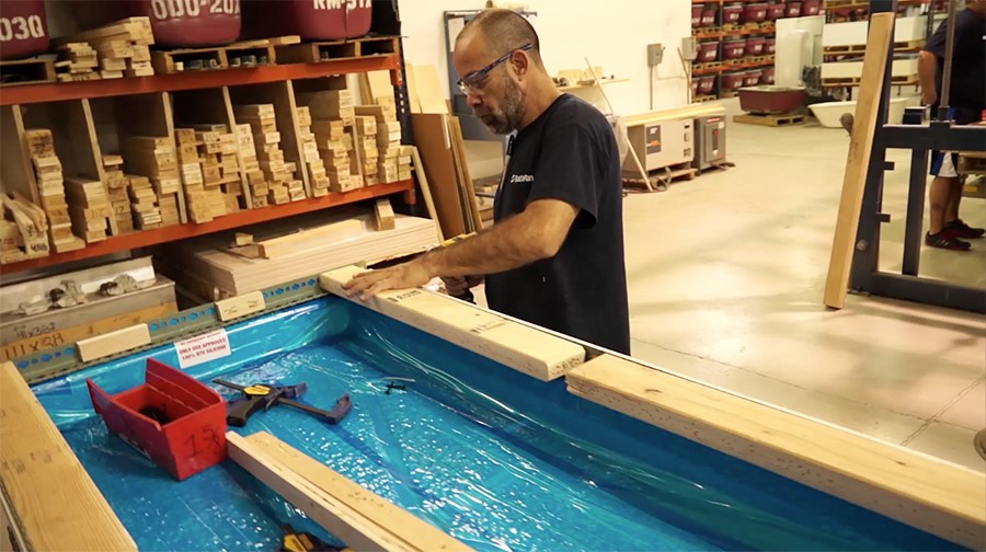 A man in a workshop fits trim boards atop a blue-lined frame. Shelves filled with lumber and tools are visible in the background.