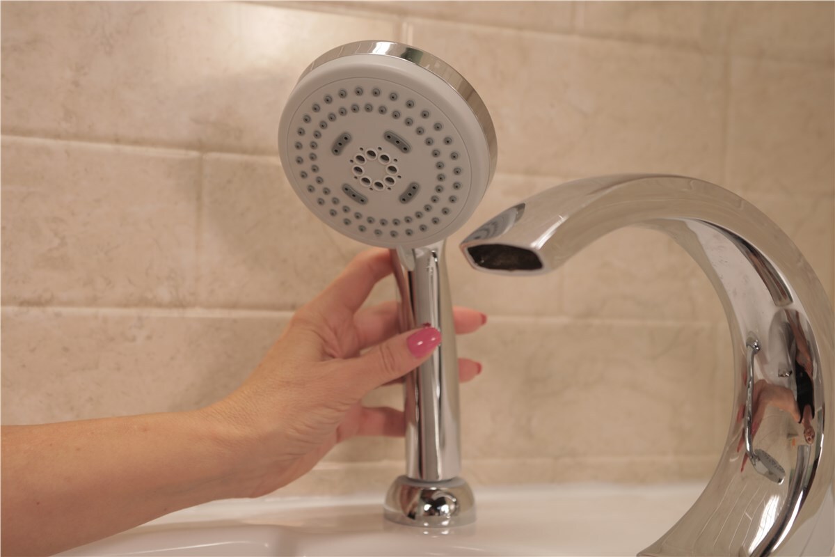 A hand holds a chrome shower fixture next to a modern faucet in a bathroom.