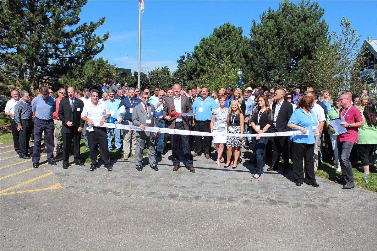 Outdoor ribbon-cutting ceremony with a large group of smiling people watching as a man in a suit holds large scissors, poised to cut the Libertyville ribbon.