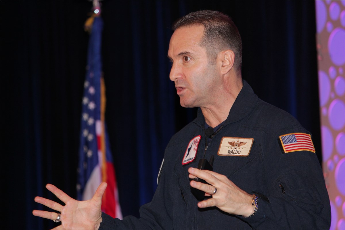 Man in a dark blue flight suit gesturing while speaking, with an American flag standing behind and to the left.