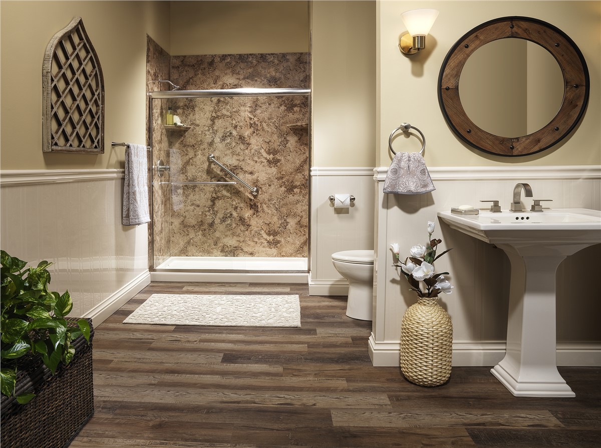 A bathroom with wood-style flooring, a pedestal sink, a round mirror, a toilet, a walk-in shower with brown marbled walls, a towel rack, wall sconce, decorative plant, and a vase with flowers.