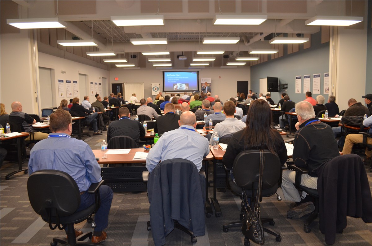 Conference room with rows of seated attendees facing a presenter on stage and a projection screen displaying a presentation.