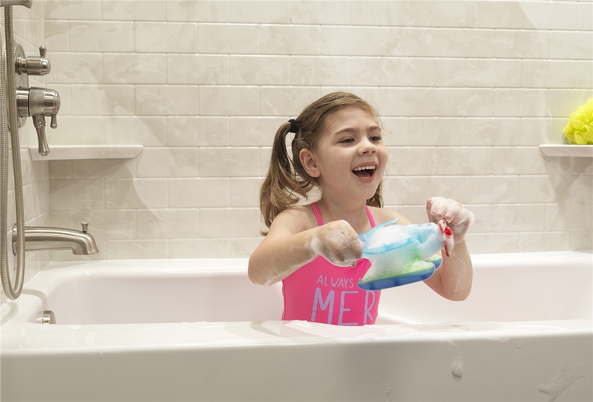 Smiling young girl in a pink swimsuit sits in a white bathtub holding a blue and green toy filled with bath bubbles.