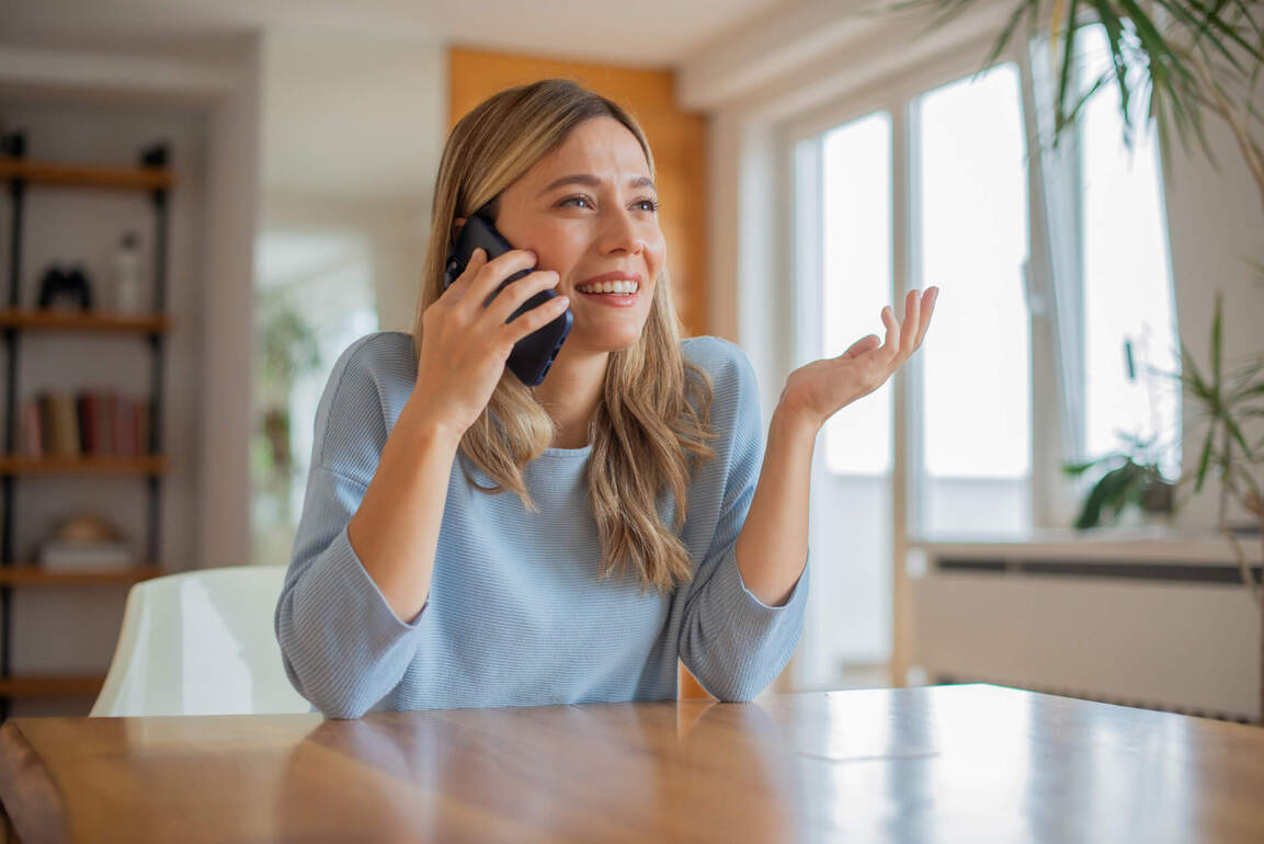 Woman sitting at a table indoors, smiling and gesturing while talking on a smartphone.