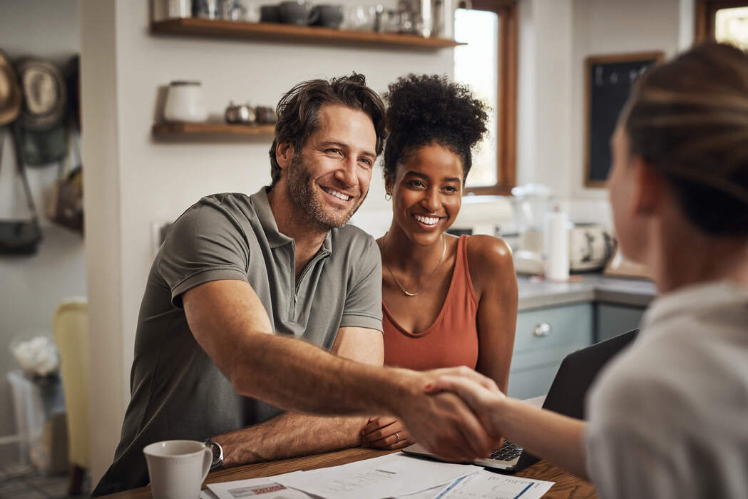 A man and woman sit at a table smiling as the man shakes hands with another person across from them, with paperwork and a pen on the table.