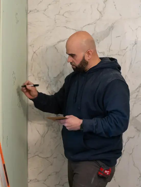 A bald man with a beard, wearing a dark hoodie, writes with a black marker on a wall while holding a clipboard.