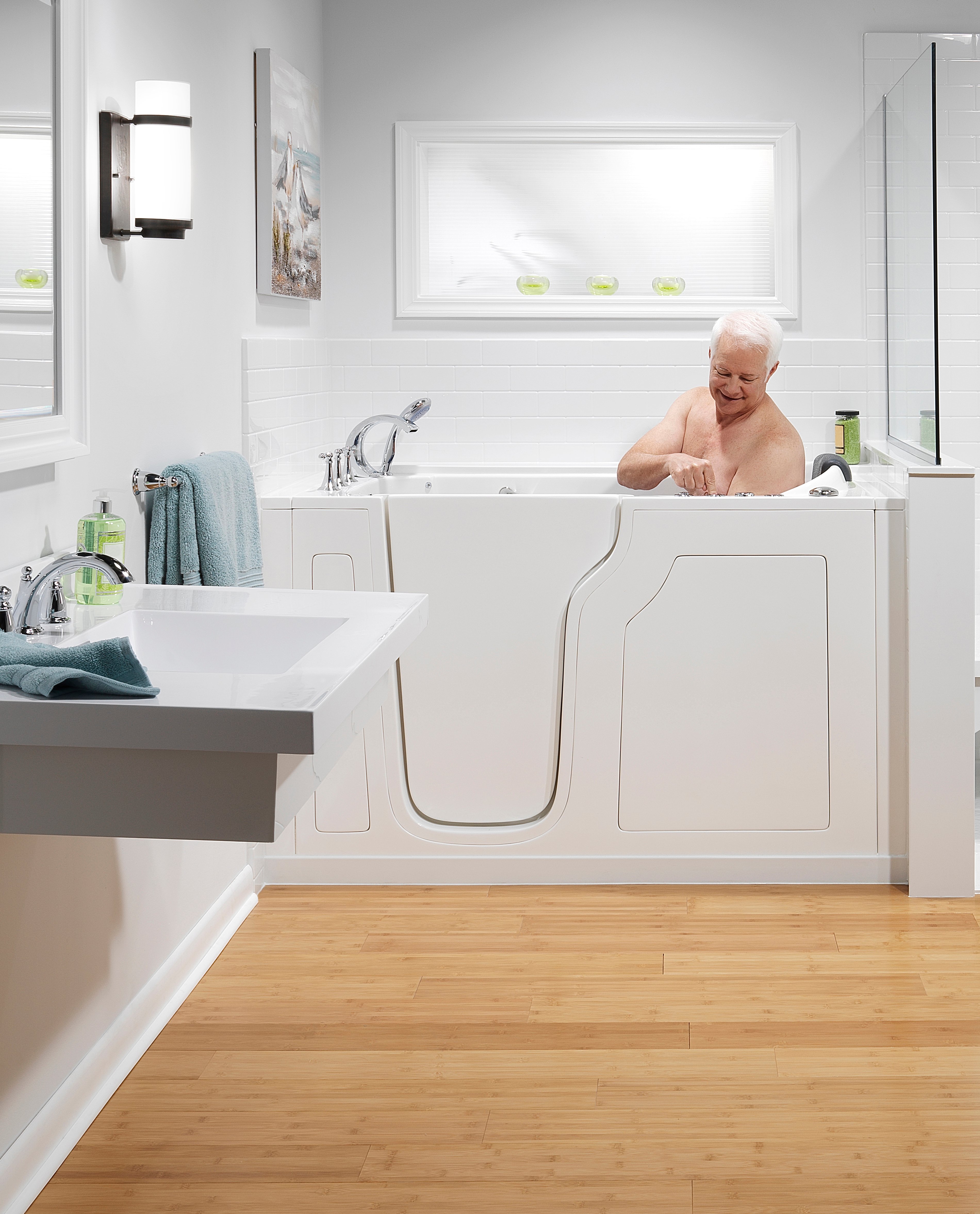 An elderly man sits in a walk-in bathtub, smiling and touching the jet controls, in a white-tiled and wood-floored bathroom.