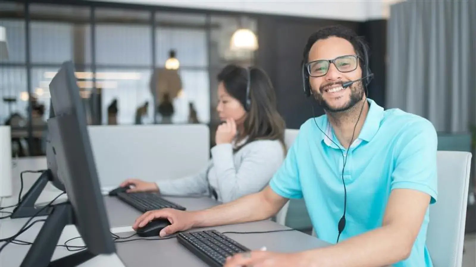 Smiling customer service representative wearing a headset while working at a computer, with a female colleague in the background.