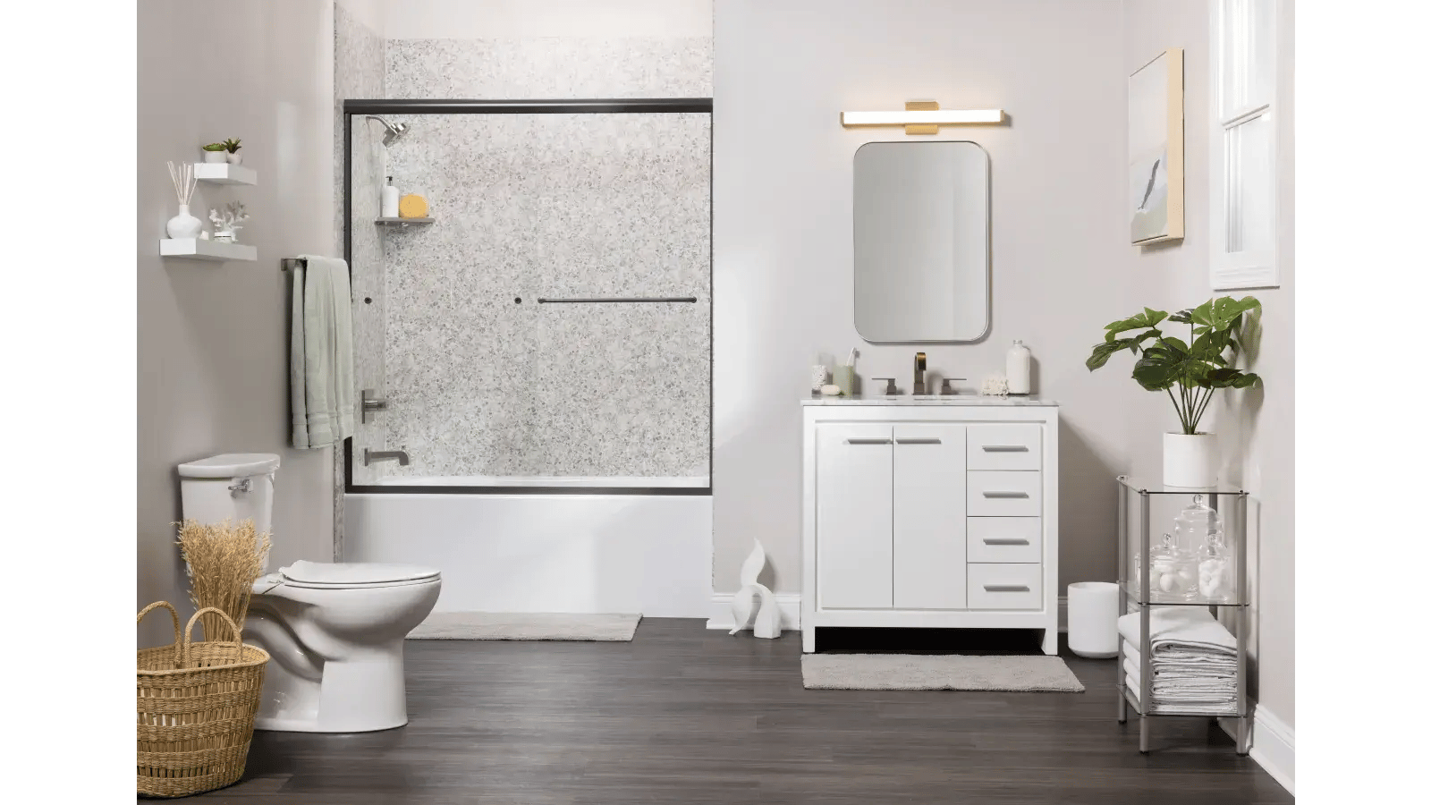 Bathroom interior with a white vanity, framed mirror with lighting, bathtub with sliding glass door and gray speckled surround, toilet, dark wood-look flooring, and decorative elements.