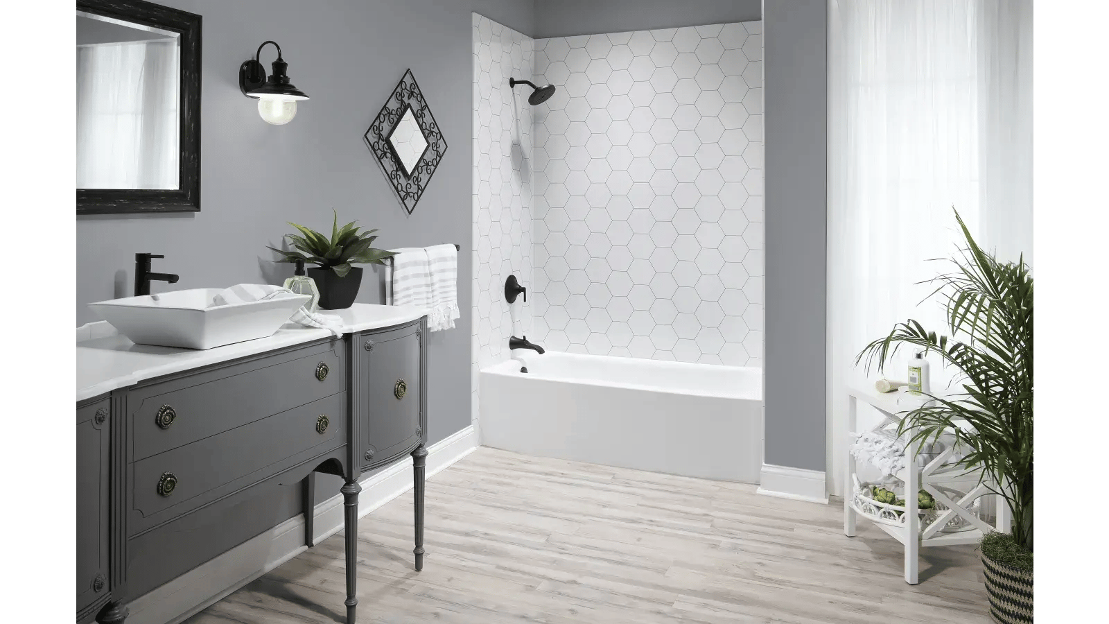 Bathroom featuring a gray vanity with a white countertop and rectangular vessel sink, white hexagon tile surrounding a white bathtub with black hardware, a black framed mirror, and light colored plank flooring.