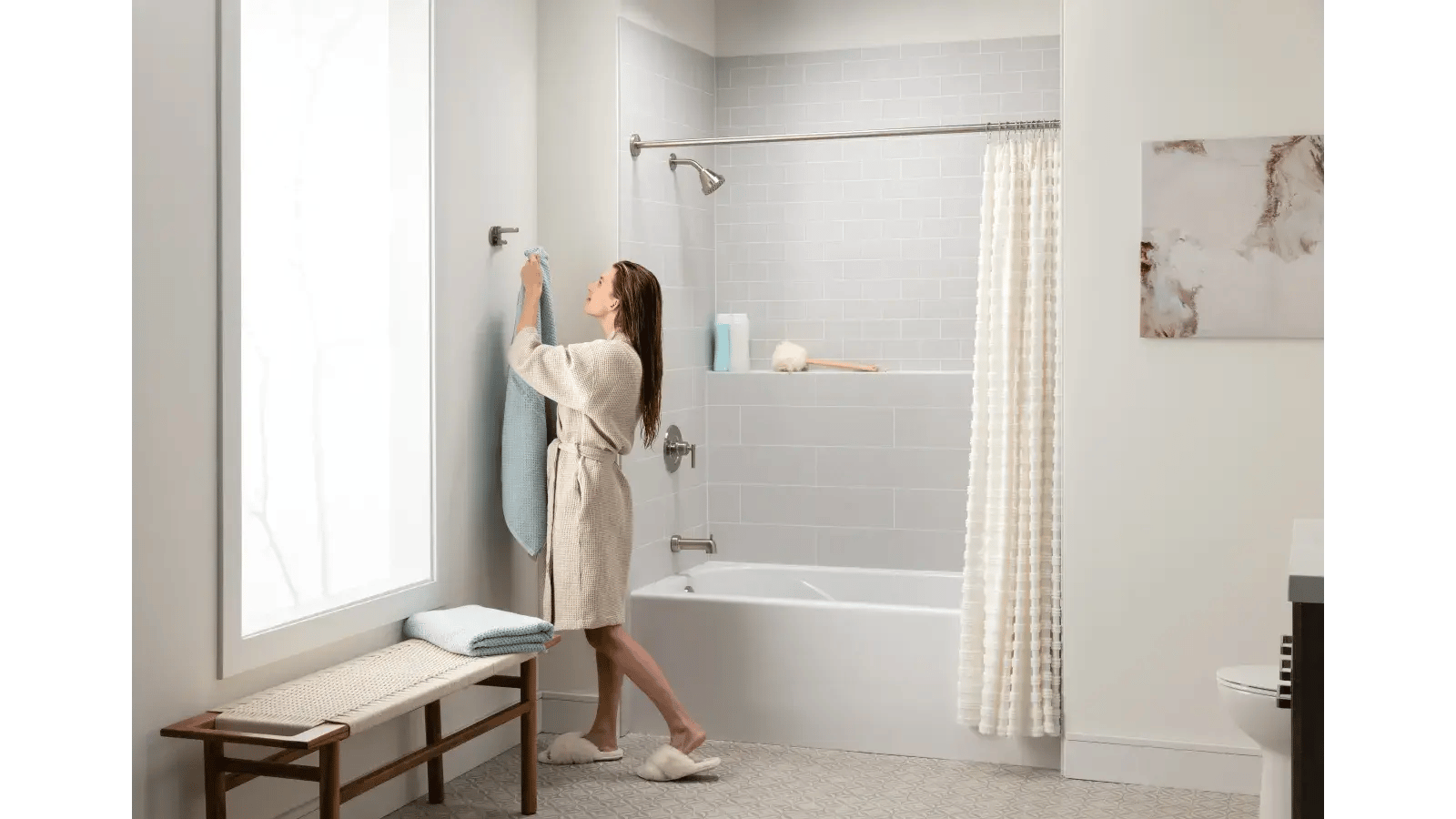 A woman in a bathrobe hangs a towel in a clean, modern bathroom with gray and white decor.