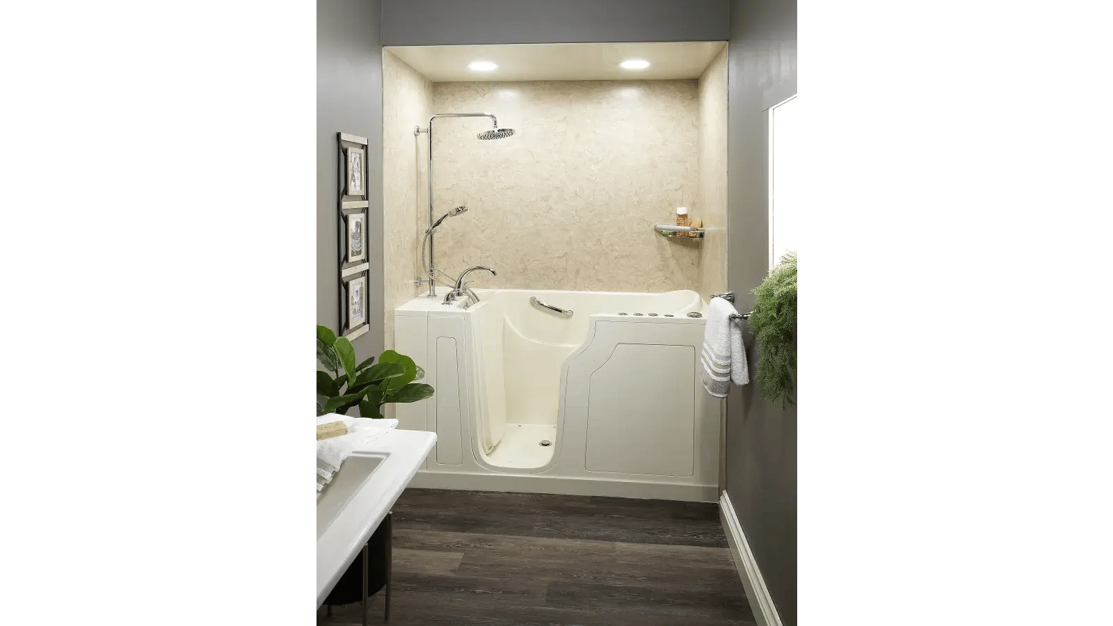 Interior bathroom space featuring a walk-in bathtub with a door, mounted handheld shower head, a shelf holding soap items, brown wood flooring, and a white sink counter.