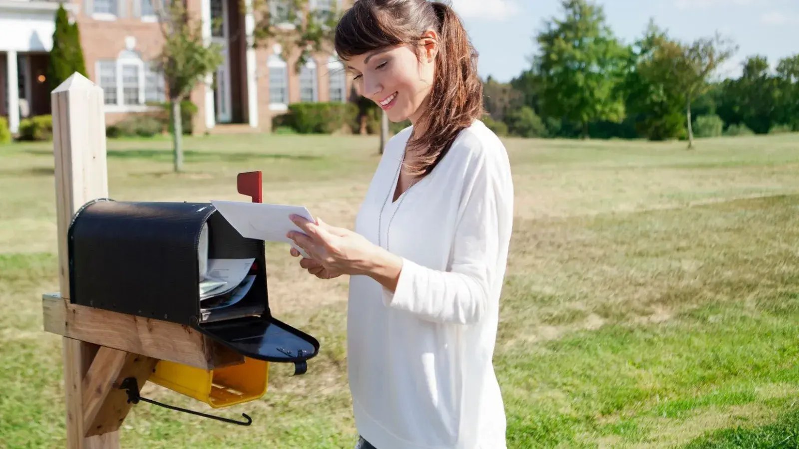 Woman smiling and placing a white envelope into an open black mailbox; a brick home and green trees are visible in the background.