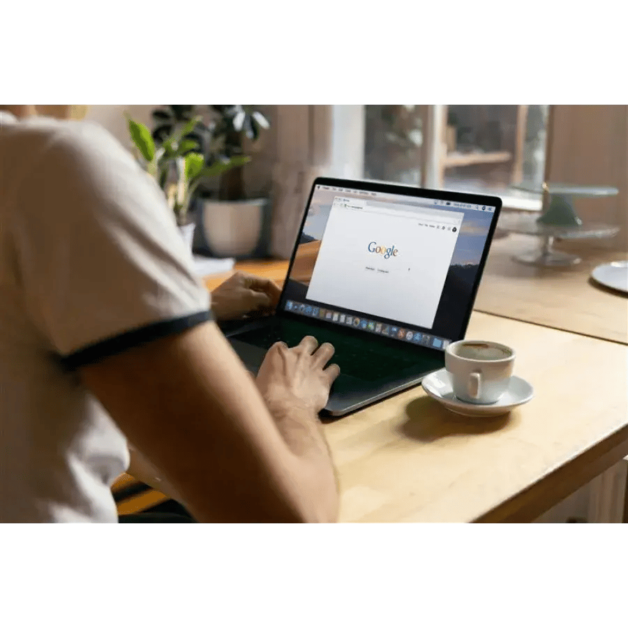 Person using a laptop displaying the Google search engine homepage, with a cup of coffee to the right.