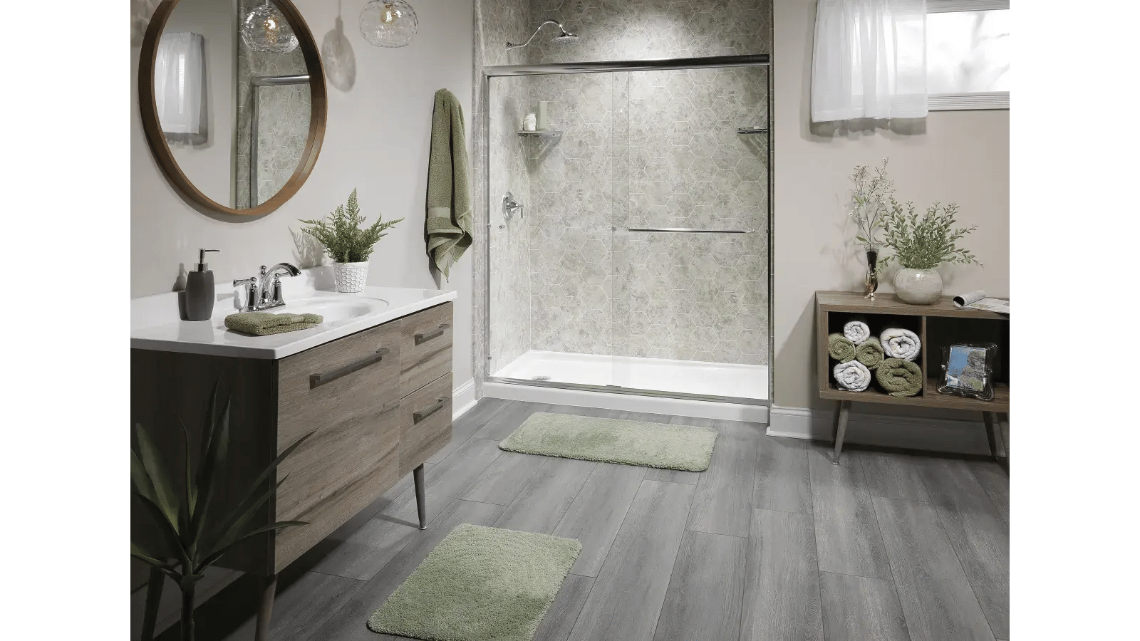 Bathroom featuring a sliding glass door shower with beige hexagonal tiling, a wooden vanity with a white countertop, a round wooden-trimmed mirror, gray wood-look flooring, and a wooden side table under a window.