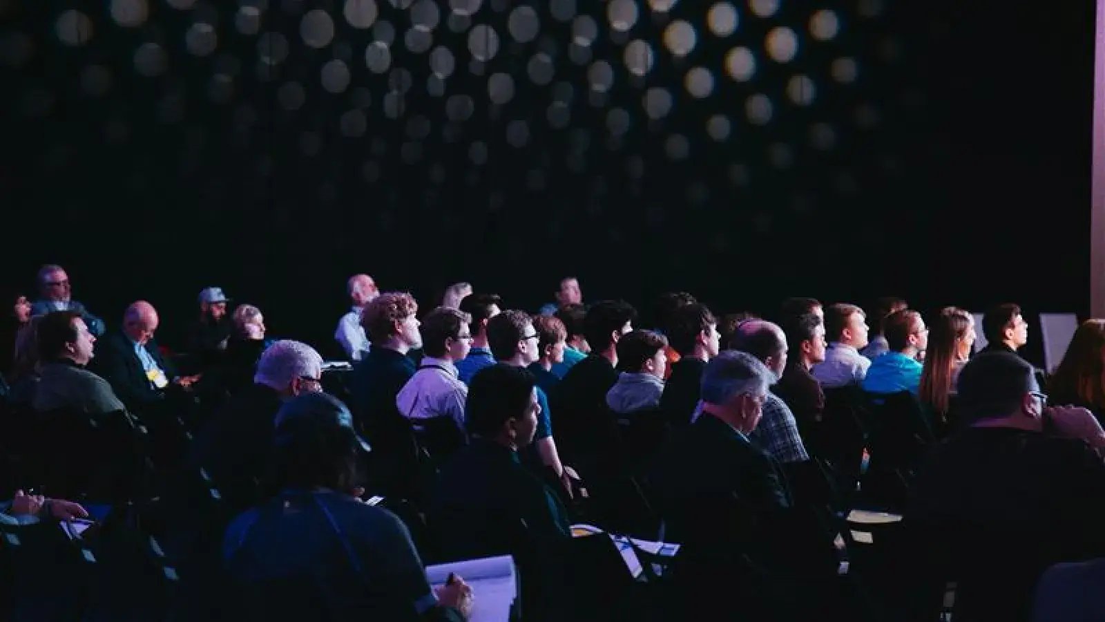 Audience members seated in a dimly lit auditorium facing forward.