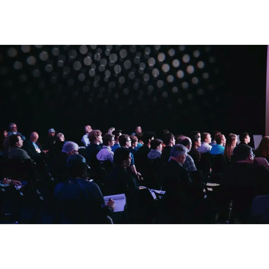 Audience seated in a dimly lit auditorium listening to a presentation; patterned lighting adorns the back wall.