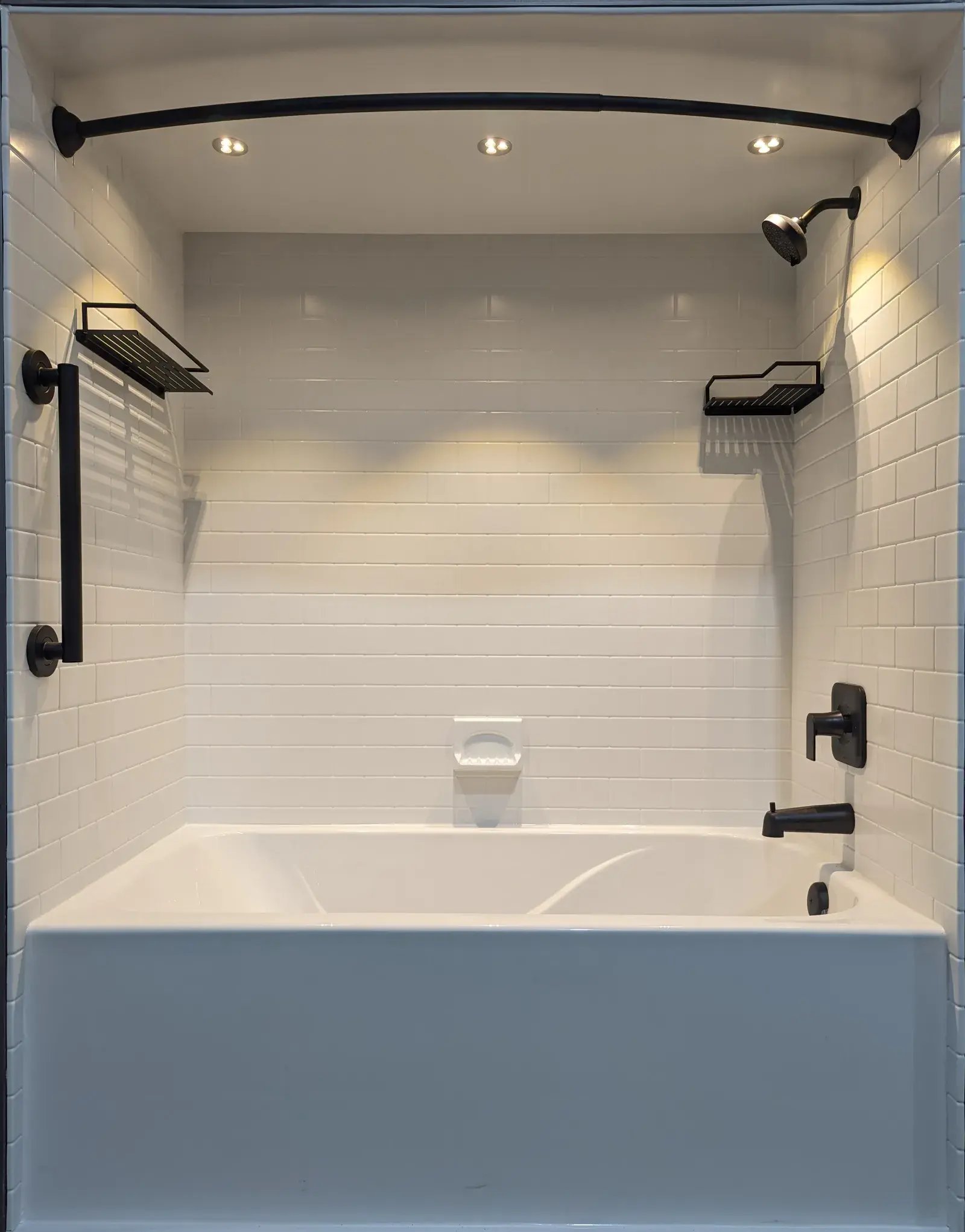 Full-Size DisplayA white bathtub with white tiled walls inside a bathroom. Black metal fixtures include shower head, faucet, handrail, and storage shelves. Recessed lighting illuminates the shower area. Examples