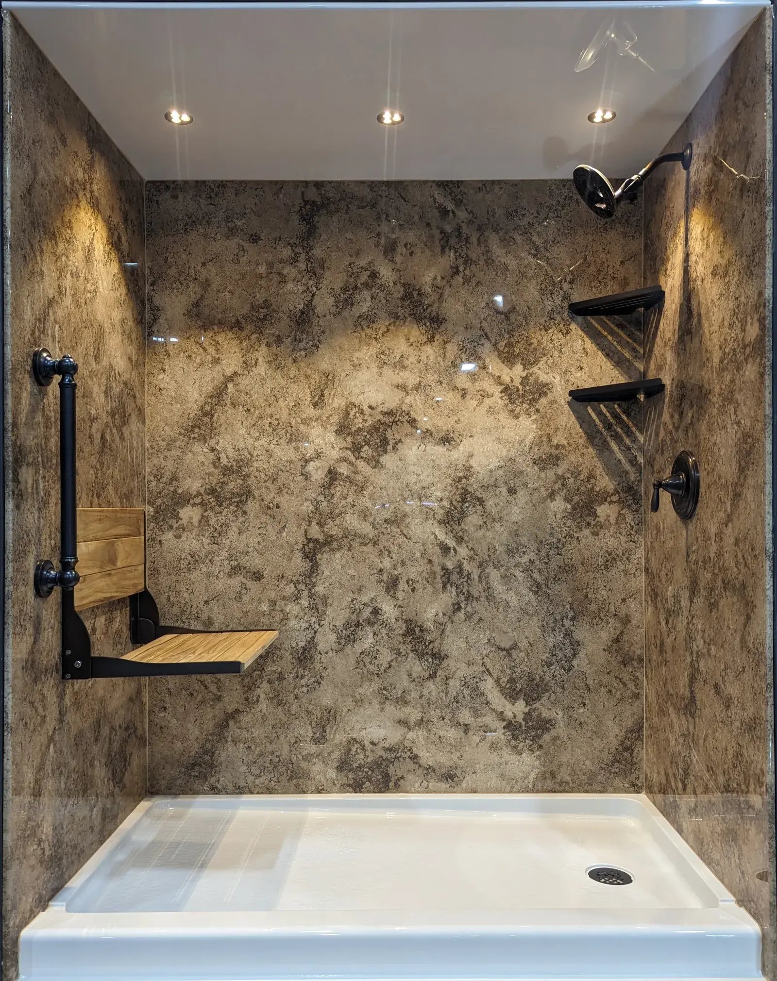 Accessible shower stall featuring a white shower pan and mottled brown wall, with folding wooden seat, overhead shower head, corner shelves and recessed lighting.