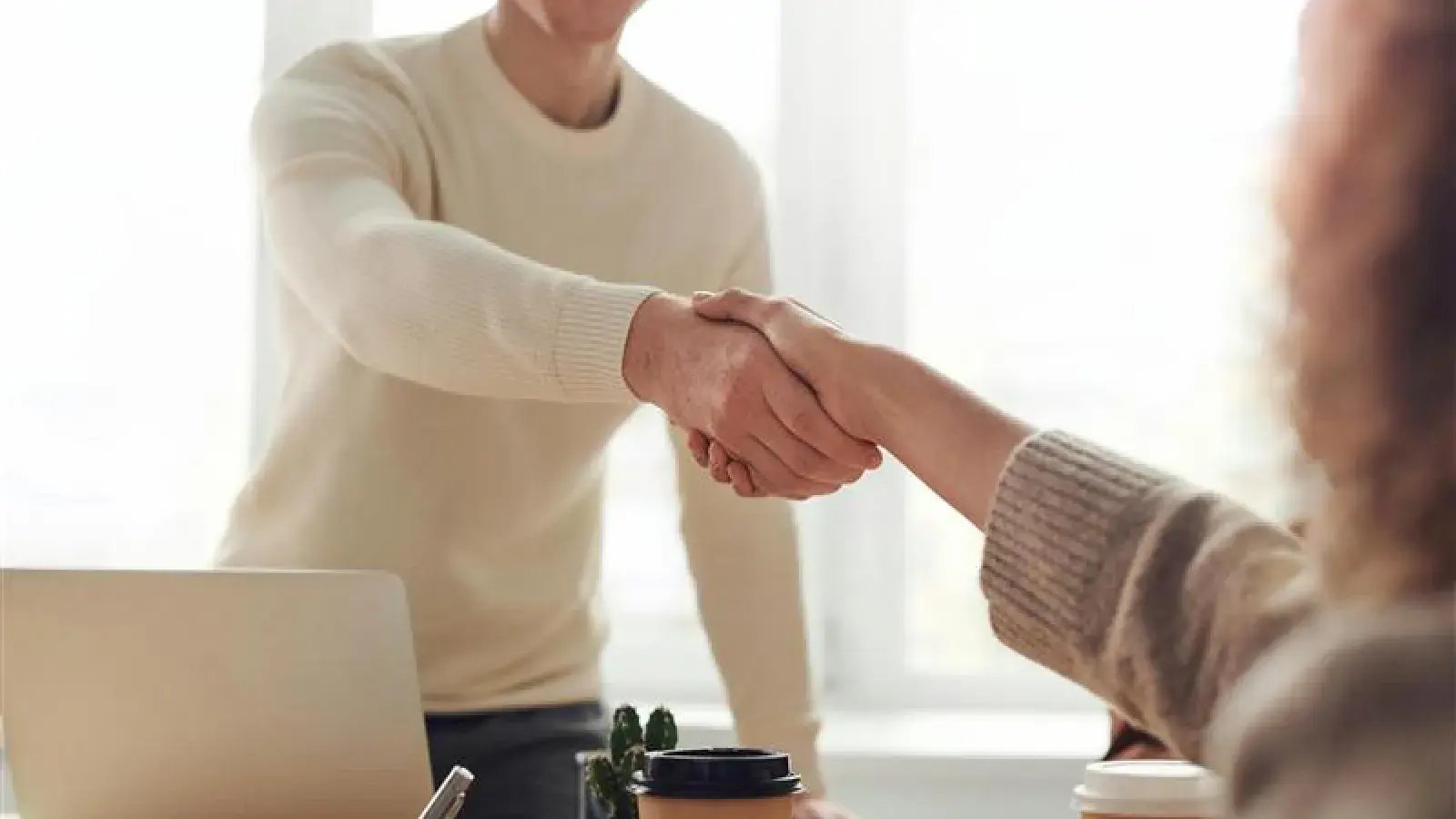 Two people shaking hands, in an office setting with a laptop, coffee cups, and a plant visible on the desk.