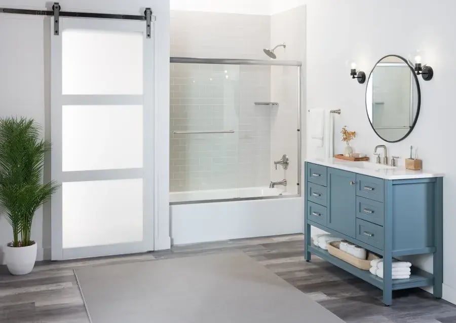 Modern bathroom with a sliding frosted glass door, a bathtub with shower, blue vanity with dual sinks, round mirror, and a potted plant on wood-style flooring.