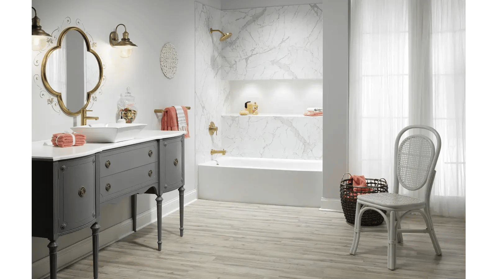 Bathroom with a grey vanity topped with a white countertop and a vessel sink with gold fixtures, a mirror with a gold frame surrounded by a decorative design, a marble-patterned shower and wall, and a white chair with a cane seat and back next to a dark woven basket.