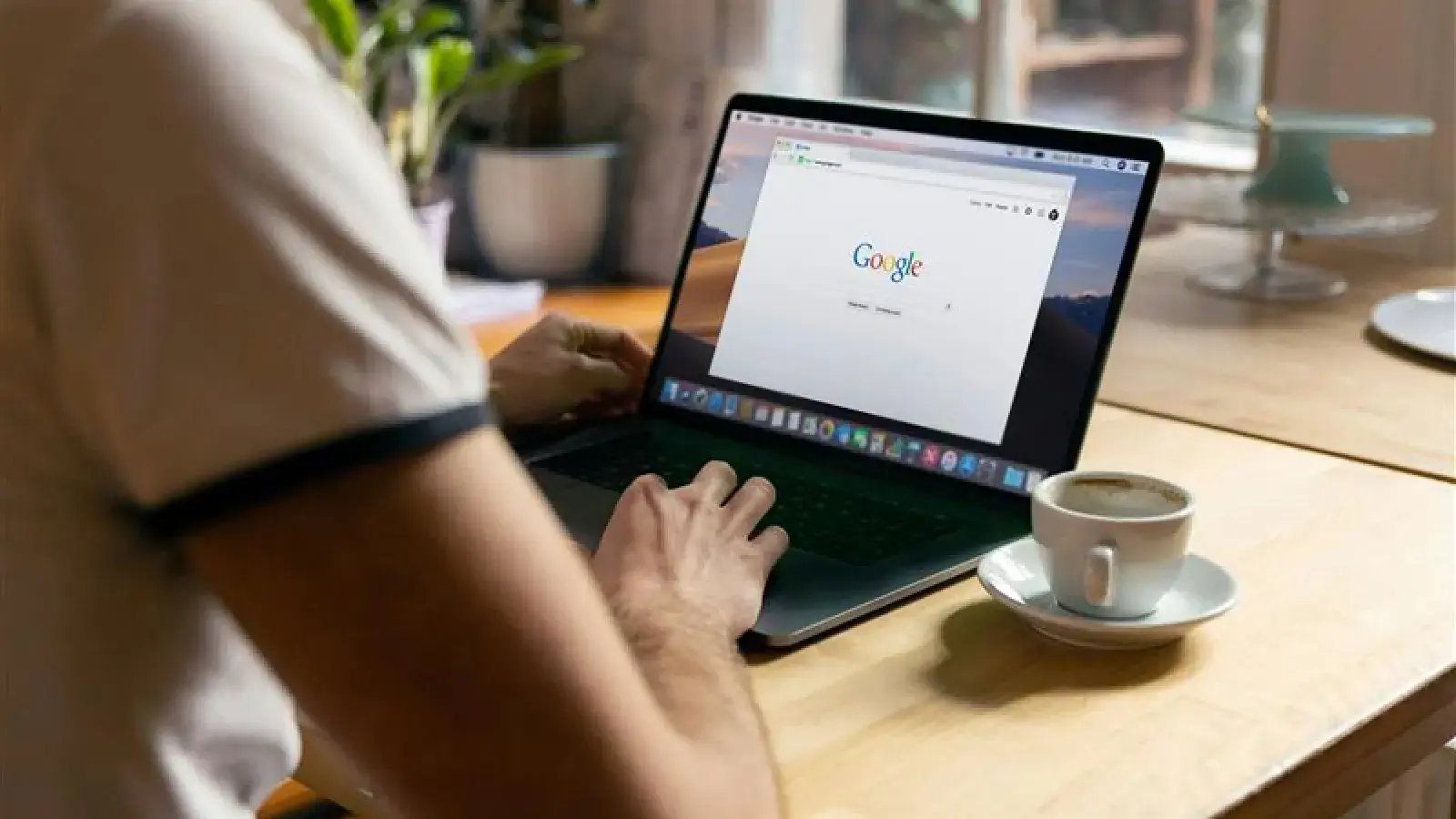 Person using a laptop displaying the Google search engine homepage, with a cup of coffee to the right.