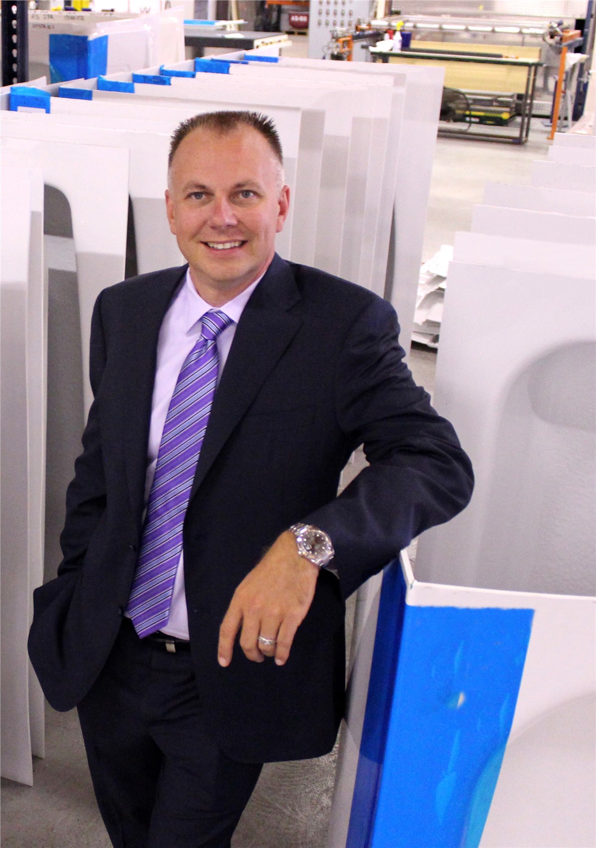 Smiling man in a suit standing in a factory surrounded by white fabricated building components.