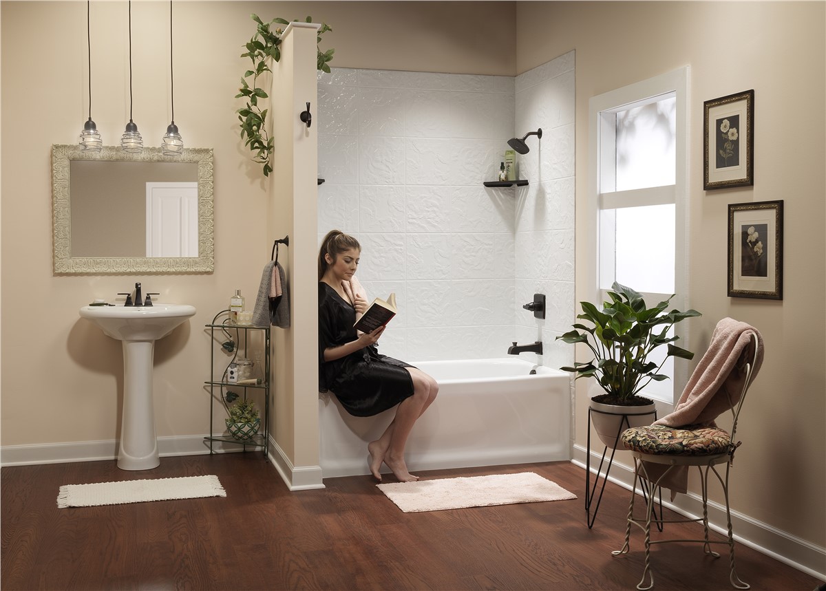 Bathroom with pedestal sink, framed art, and a woman sitting on the edge of a bathtub reading a book.