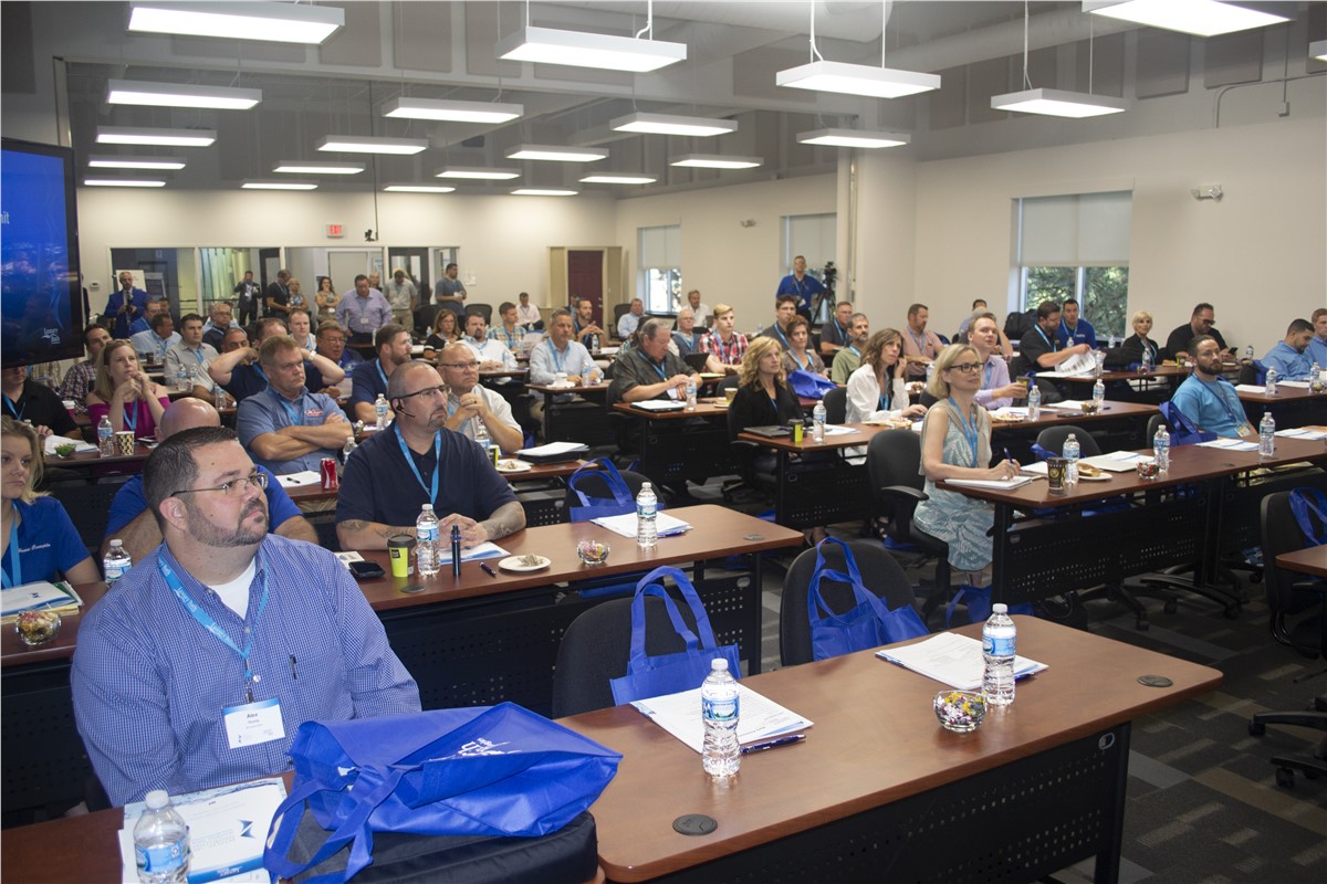 Large group of people seated at desks in a conference room, attentively watching a presentation.