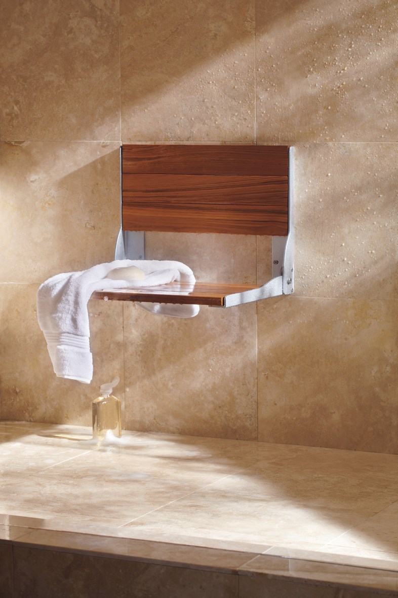 Wall-mounted shower seat with a wooden slatted surface and metal support brackets, folded down within a beige-tiled shower. A towel and bar of soap sit on the seat, with a dispenser-style bottle underneath.