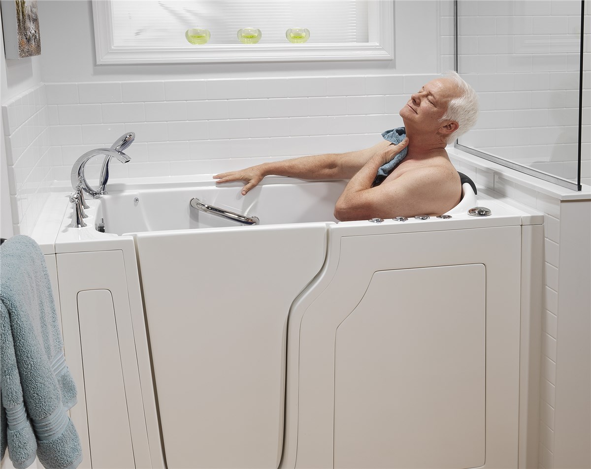An older white-haired man sits relaxed in a white accessibility walk-in bathtub, eyes closed and head tilted back with relaxed expression. He holds a light blue towel beneath his chin. The tub features a side door and supporting grab handles.