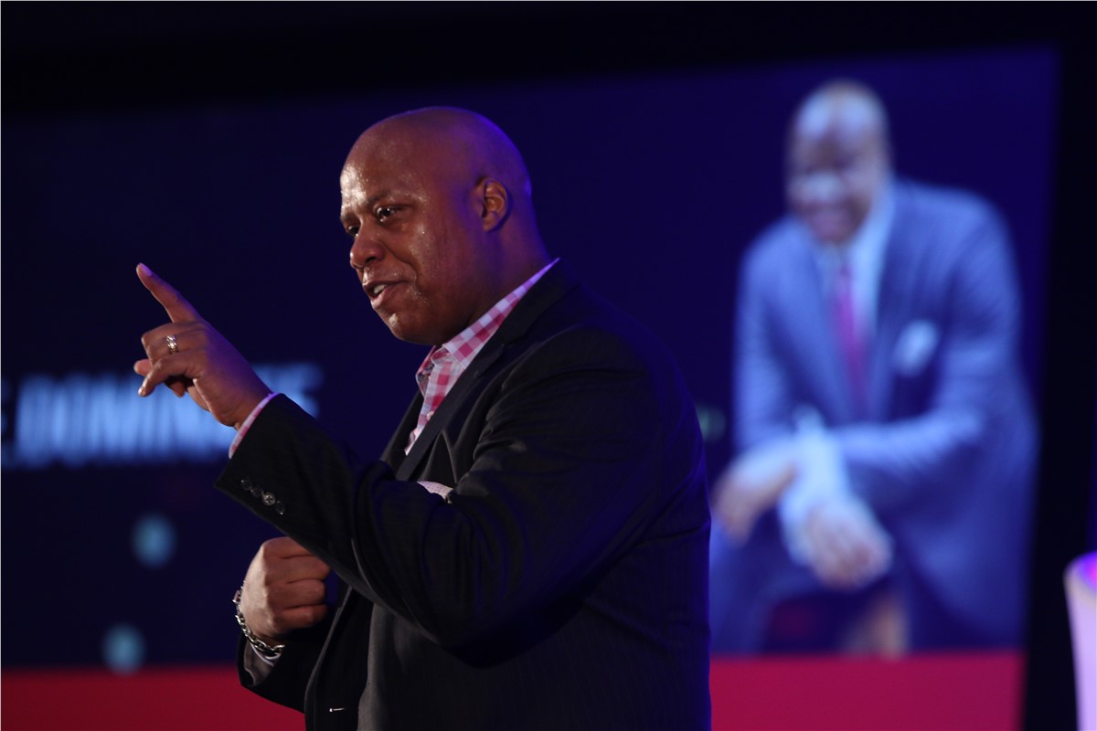 A bald man giving a presentation. The man in focus is pointing upward, wearing a pink-and-white checkered shirt and black jacket. Behind him is a large screen showing another man, in a suit, smiling. The background is dark blue.